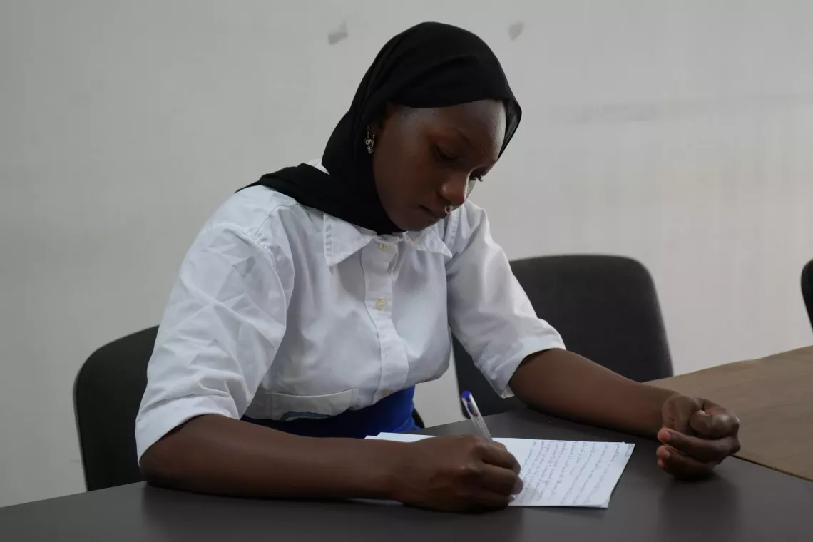 A girl in a black scarf sits at a table writing a letter in Mali. 