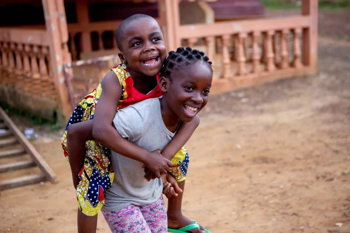 A young girl smiles as she carries a friend on her back in Cameroon.
