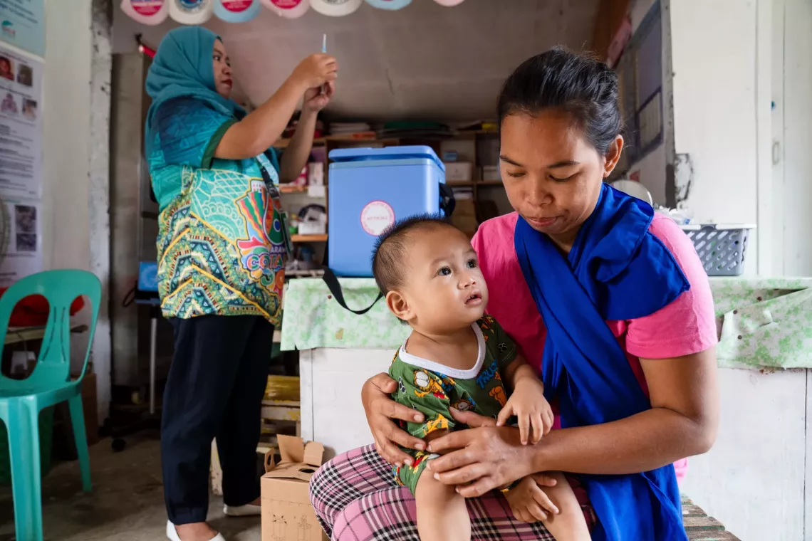 A child sits on the lap of their mother while a health care worker prepares a vaccine shot in the background