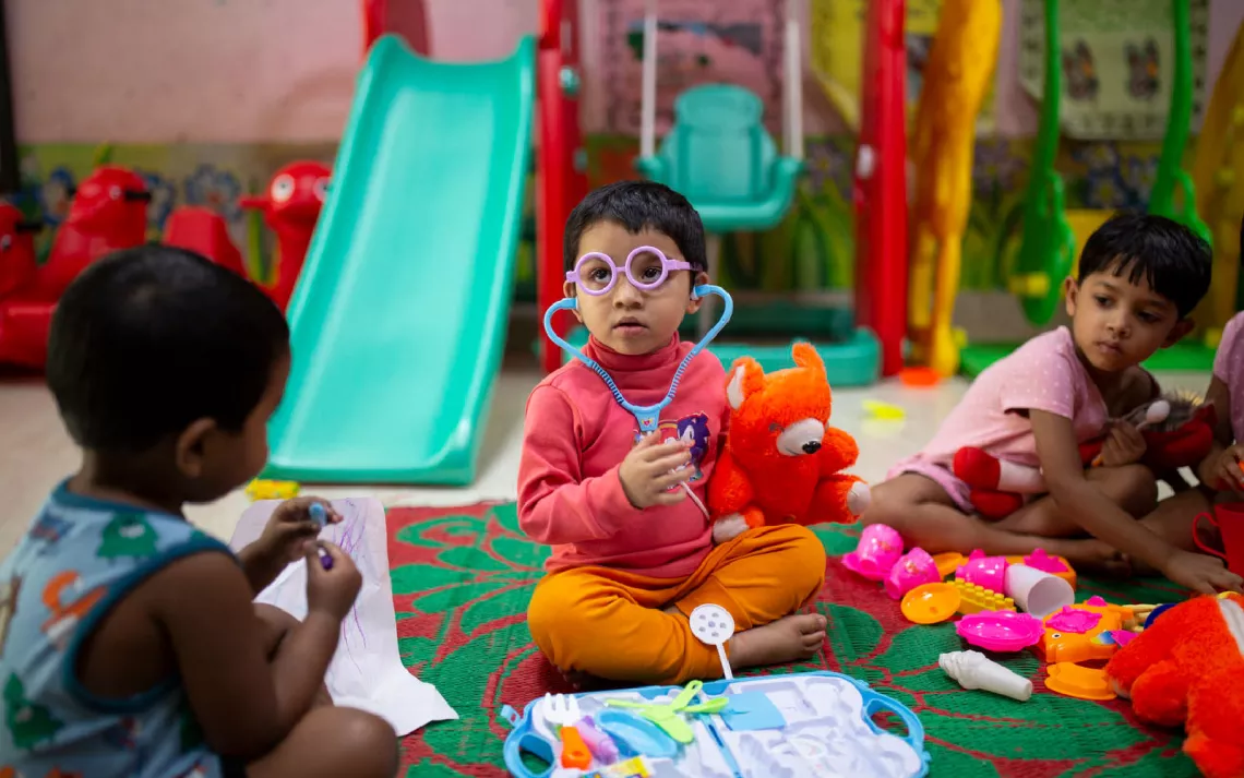 A child wearing a toy medical kit sits on the floor of an Early Childhood Devleopment Centre in Bangladesh