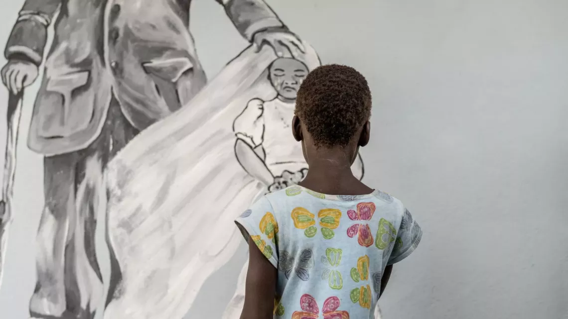 A young girl in Mozambique stands in front of a mural depicting a child in a bridal veil crying.