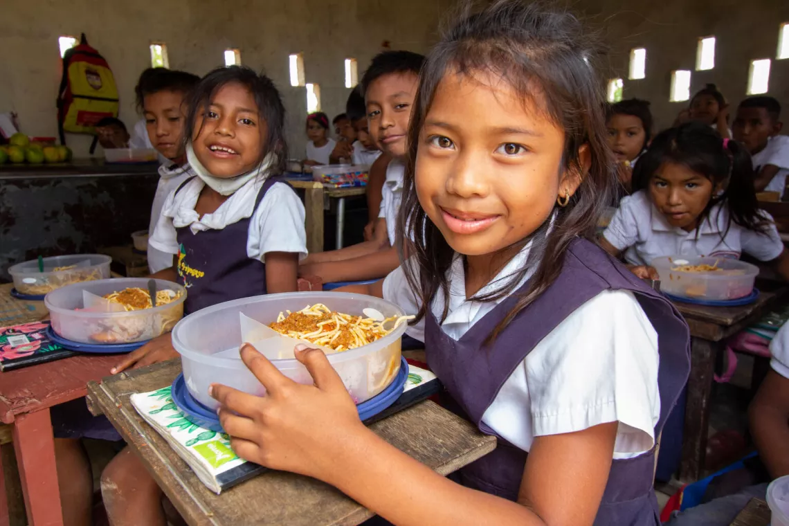 Children smile at the camera while they eat a plate of hot food at a UNICEF-supported school.