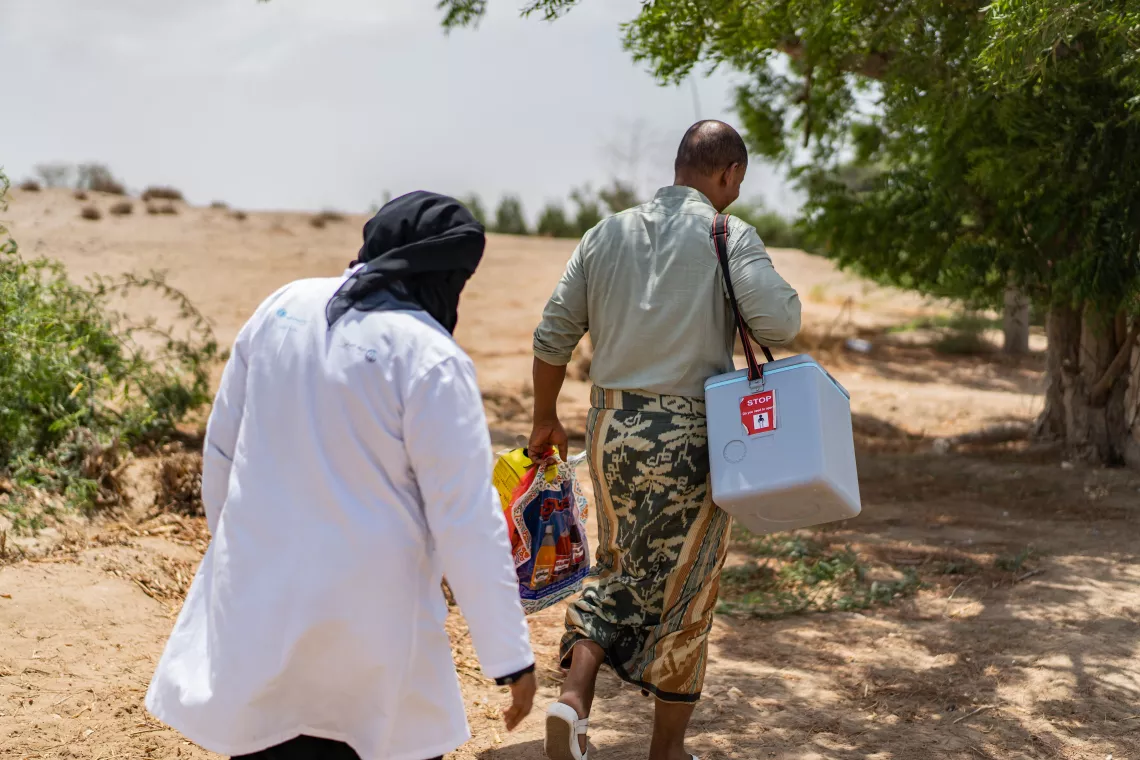 Health worker Ghada Ali Obaid, 53, walk with her husband, Ehab Faisal, 43, carrying her equipment for her at the site of a community outreach vaccination drive for children.