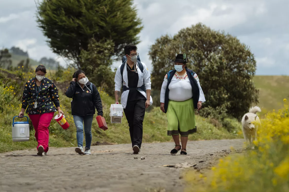 Maria, a Community Watcher walks along a road in the countryside with 2 female and a male doctor. 