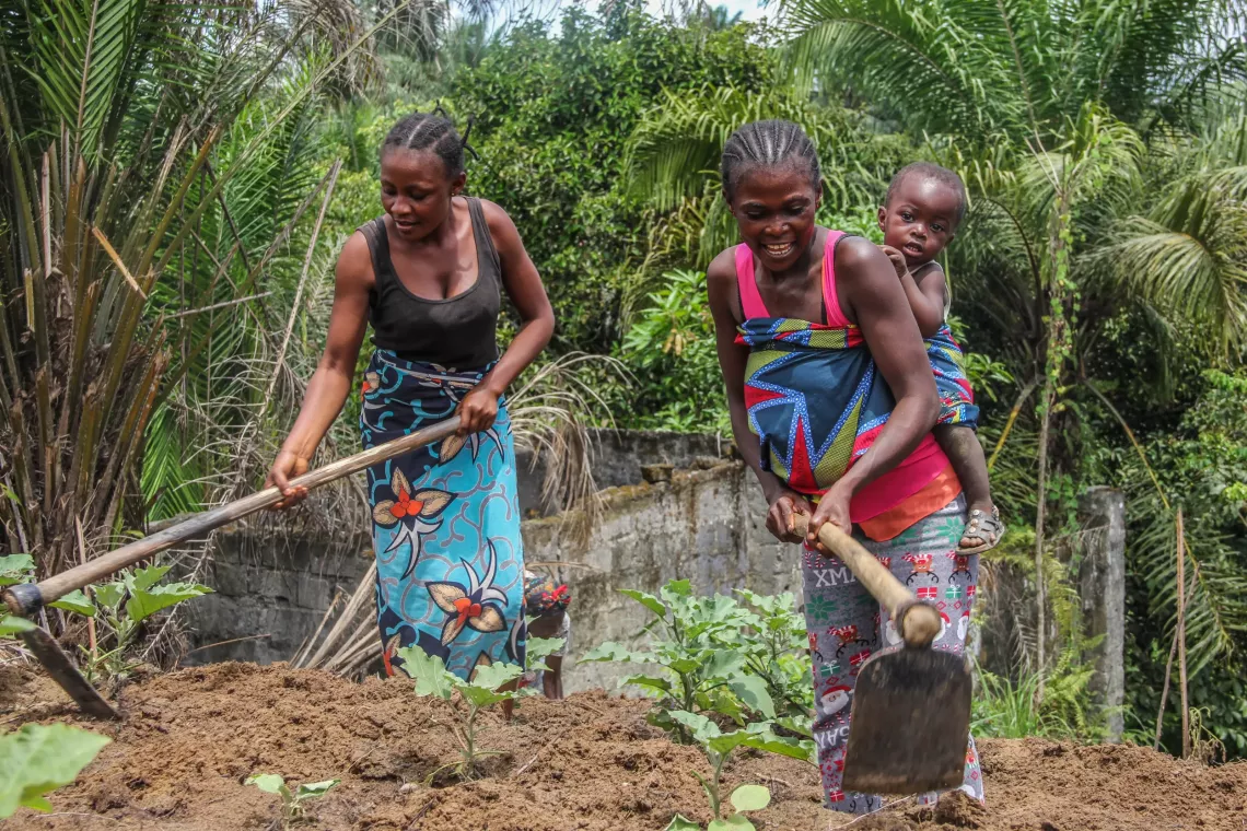 Two women ploughing a field with, one of them with a baby on her back.