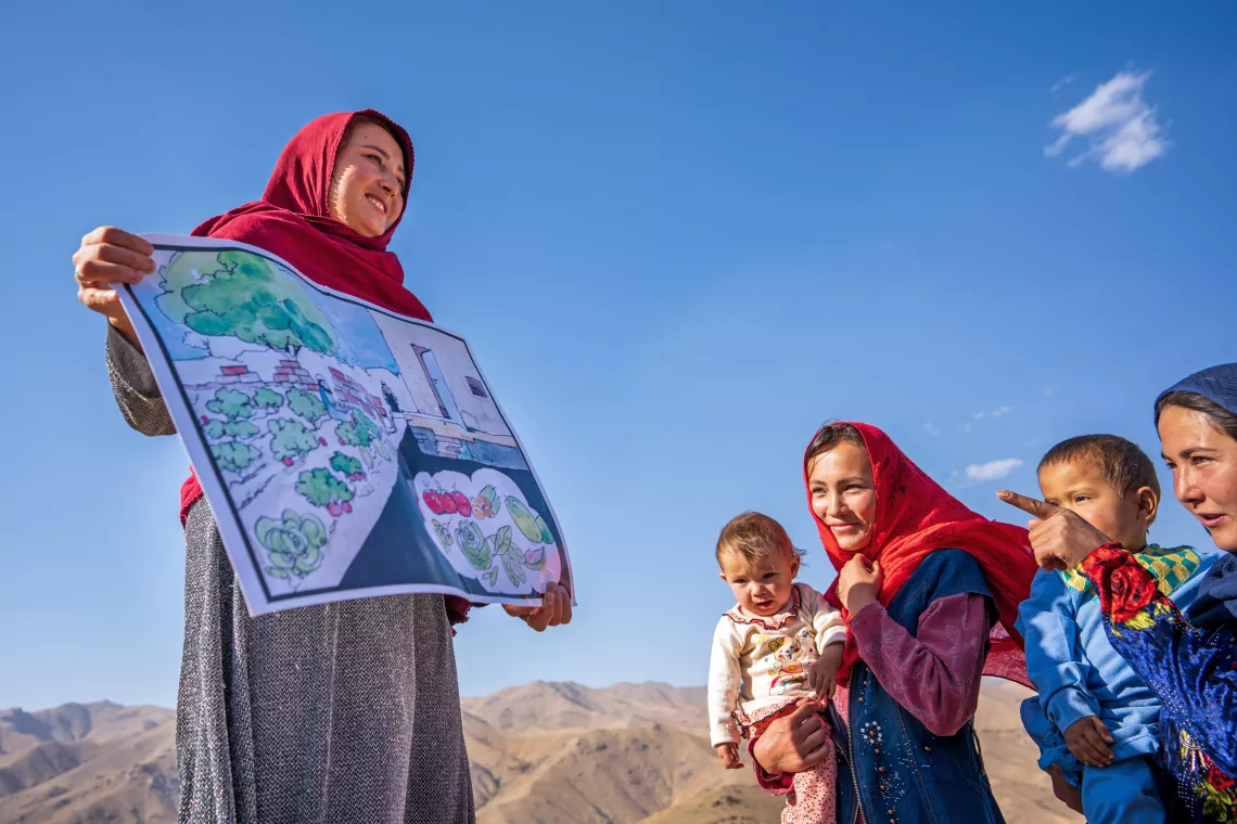 Somaya, a community health worker, holds a poster to help her educate mothers about the importance of good nutrition. They are on top of a bare hill and a woman holding a child is pointing at the poster.