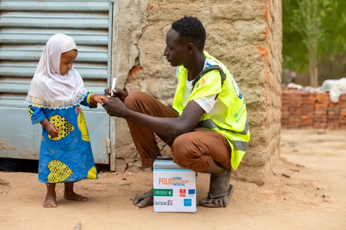Un agent de sant&eacute; en gilet r&eacute;fl&eacute;chissant vaccine un jeune enfant devant un b&acirc;timent ; une glaci&egrave;re &agrave; vaccins est pos&eacute;e au sol.