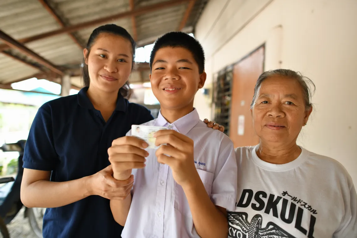 13 year old Sarm holds up his ID card smiling, while his mother and grandmother stand at his side.