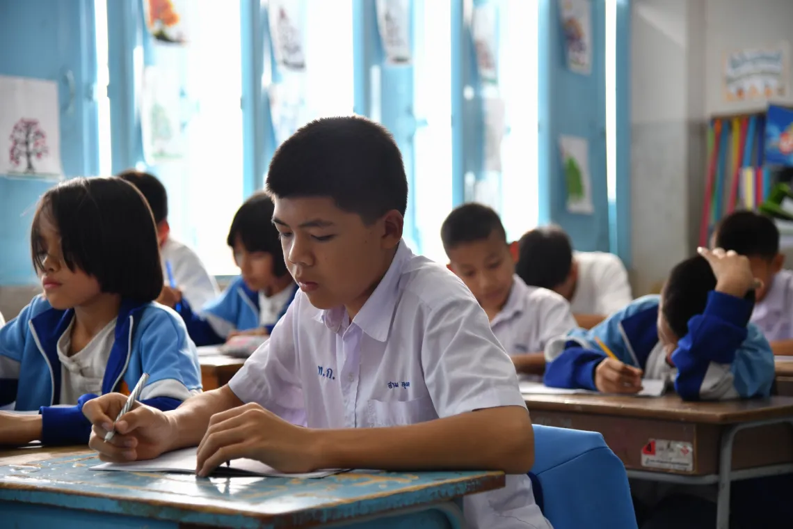 Sarm, 13, writes at his desk during class, surrounded by classmates. 