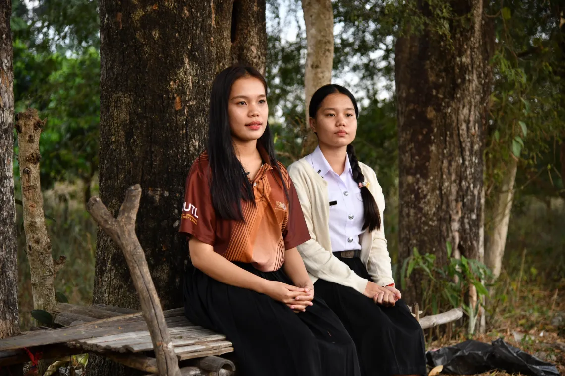 Kanokwan and Kamonwan sit together on a wooden bench beneath the trees
