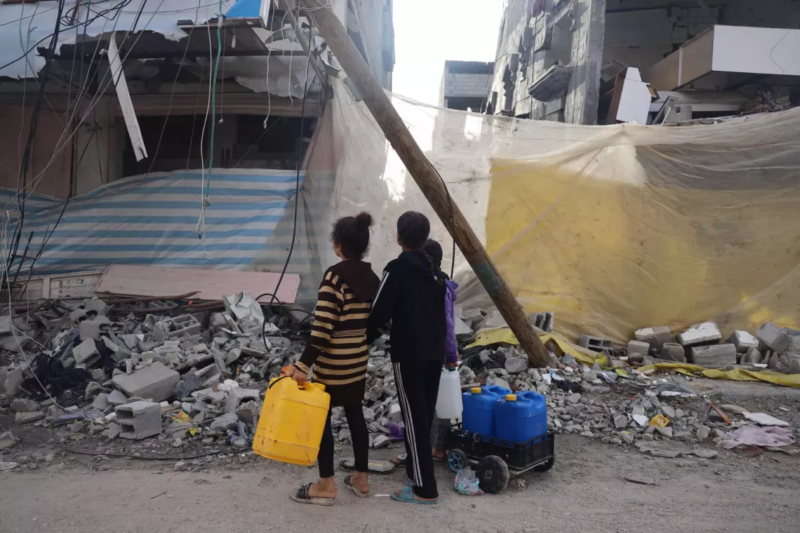 State of Palestine. Children collecting water walk past badly damaged buildings.