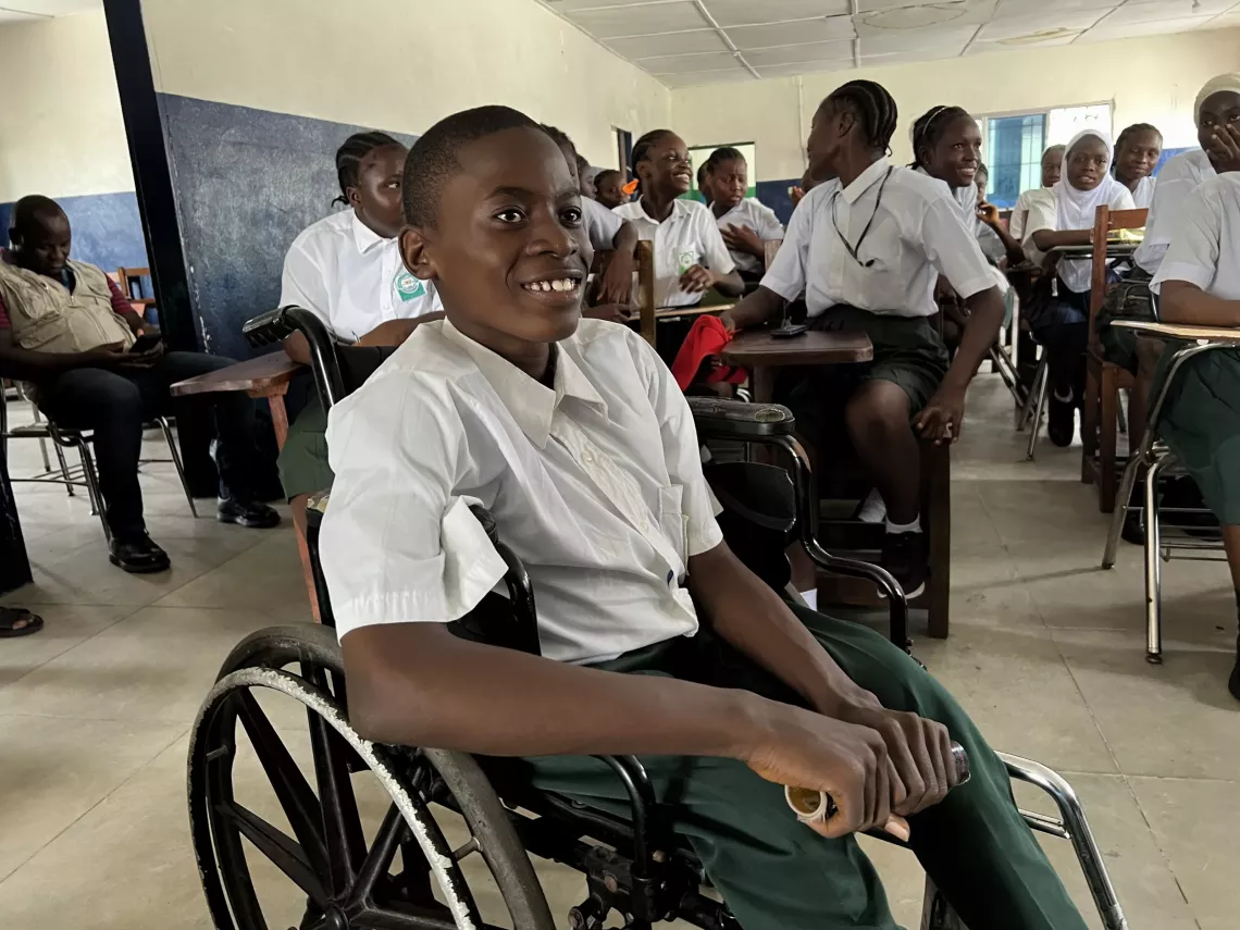 A young boy in a wheelchair smiles while seated in a classroom.