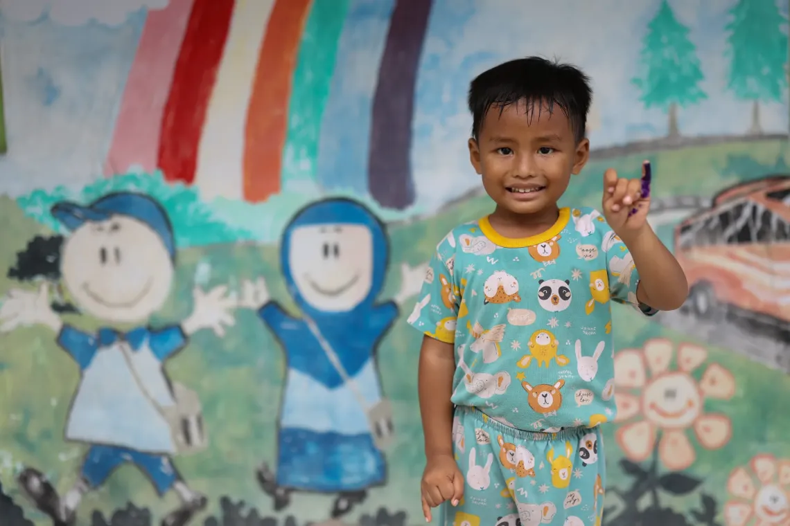 A four year old boy holds up his marked pinky finger while smiling.