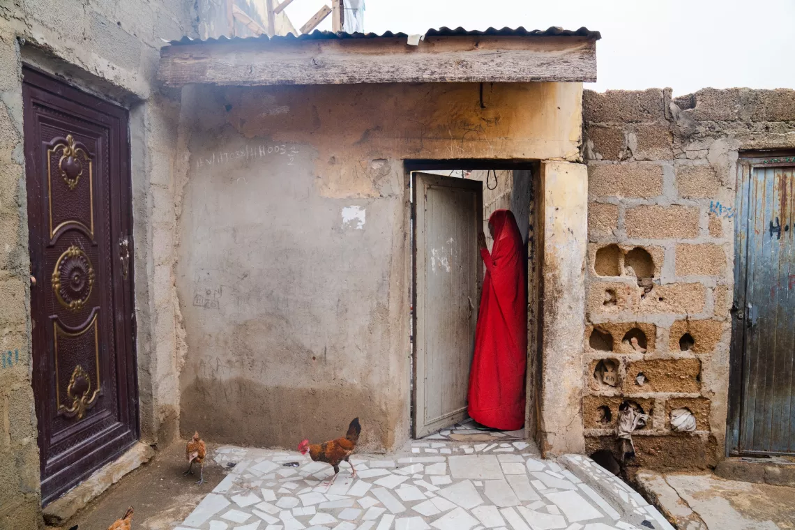Shamsiyya, dressed in red, enters a doorway in  Katsina, Nigeria.