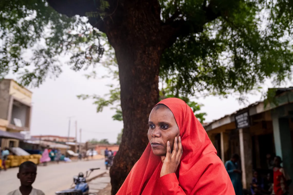 Shamsiyya stands under the shade of a tree, holding her hand to her cheek as she looks out.