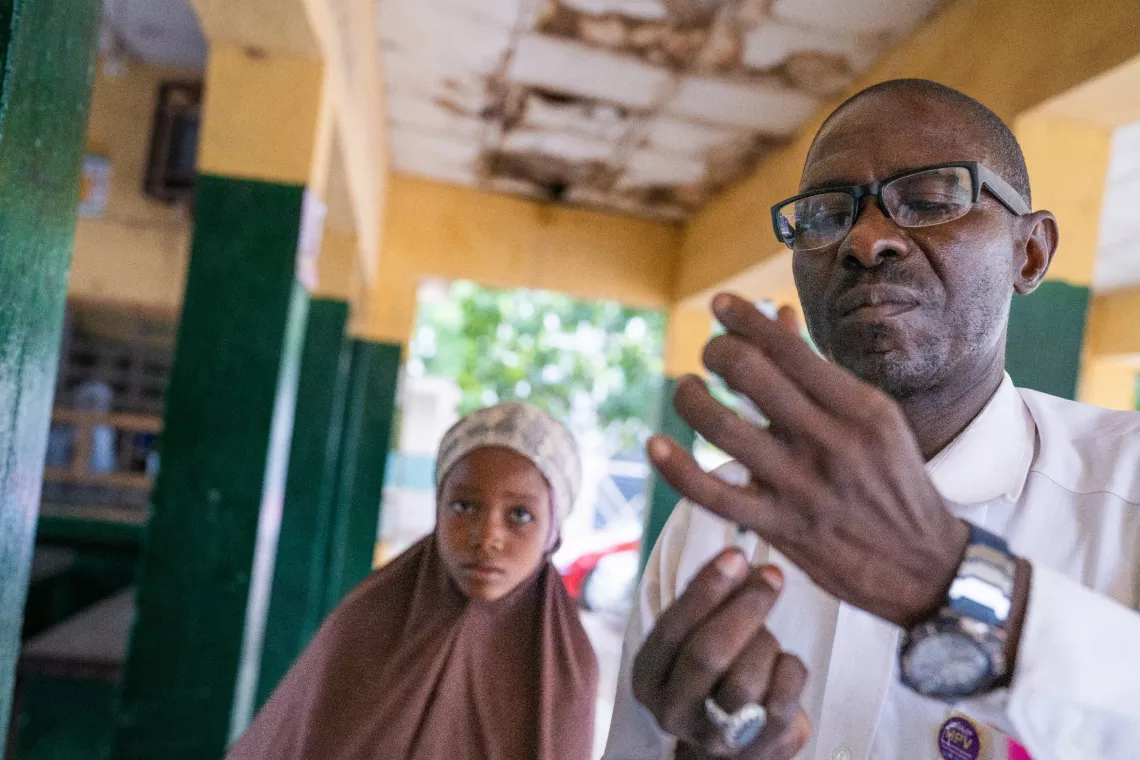 Safiyya, 10, at rear, watches as Alimudeen Mohammed, right, ward in charge at the Katsina Kofar Marusa Maternal and Child Health Clinic, prepares a human papillomavirus (HPV) vaccine.