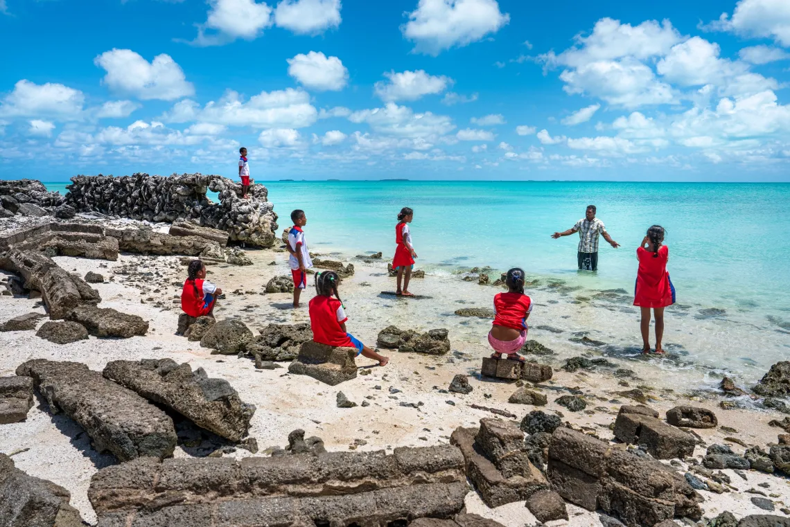 A man stands in the water, sharing stories with children as he points to where his home once stood in Tebunginako Village, which has been severely affected by erosion and rising sea levels on Abaiang Island, Kiribati