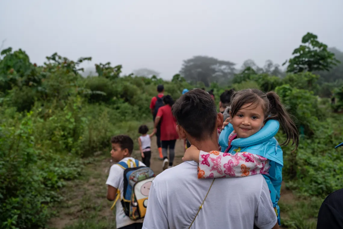 A young girl smiles while being carried by her father. Other migrant families walk ahead.