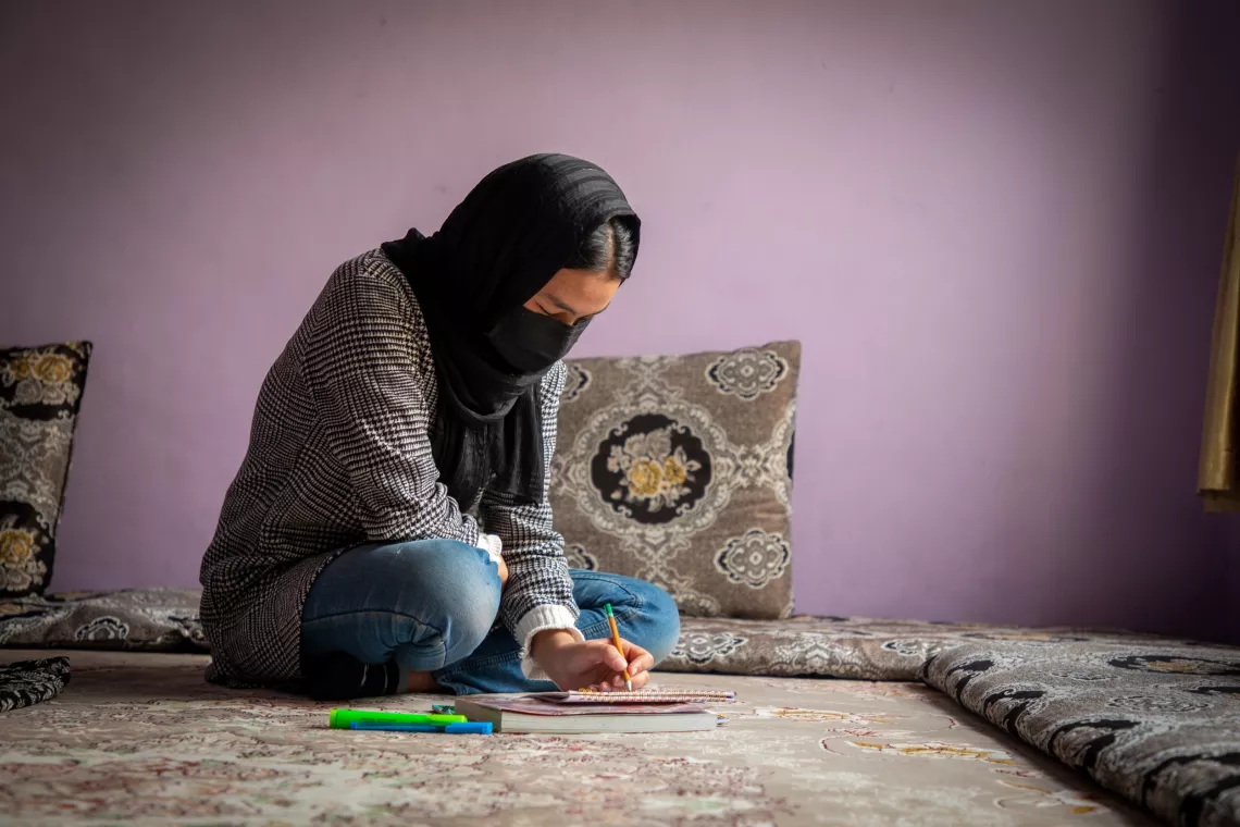 A girl sits with crossed legs while writing in book in front of her on the floor.
