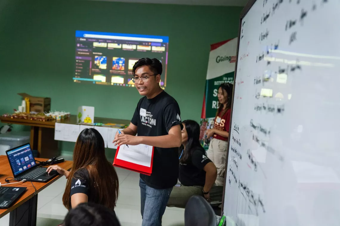 Luster Anabieza standing in front of a whiteboard addresses youth 