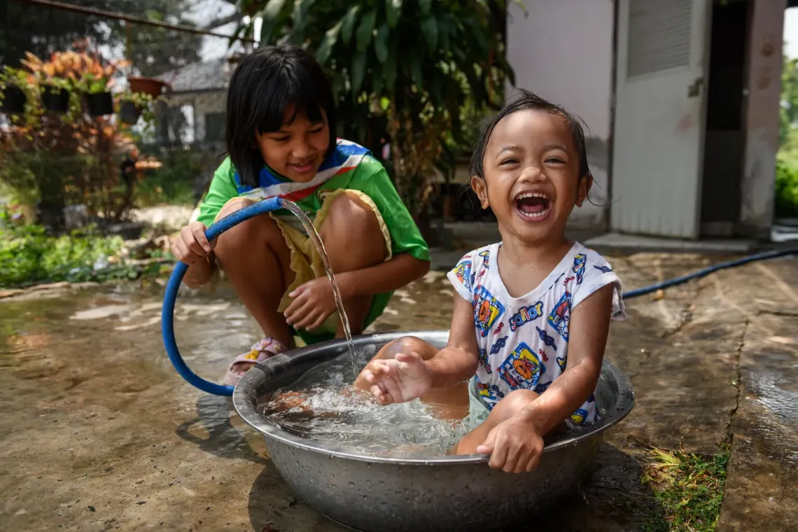 A young child sits laughing a water basin as their sibling holds a hose spouting water. 