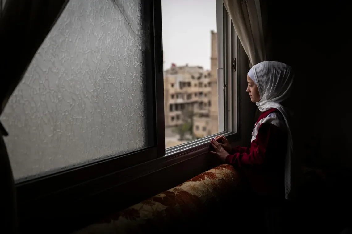 An eleven year old girl in a white head scarf looks out the window of her home in Ain Tarma, Syria.