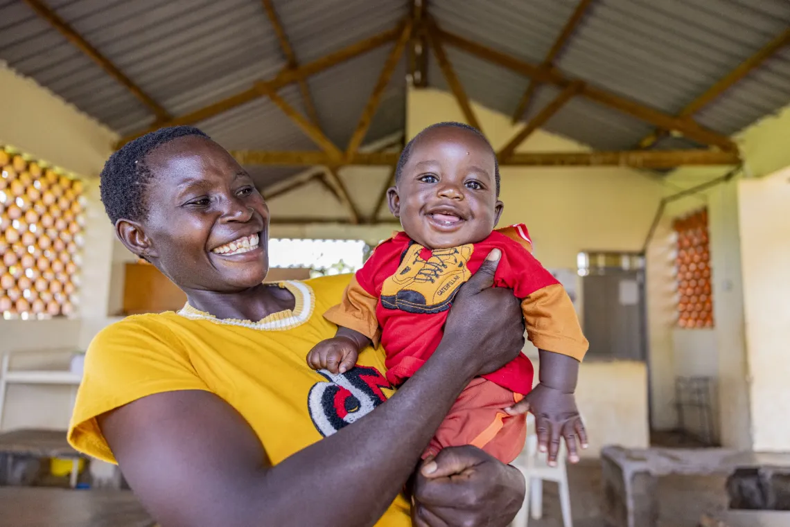 A smiling mother in a yellow shirt holds up her brightly smiling baby. 