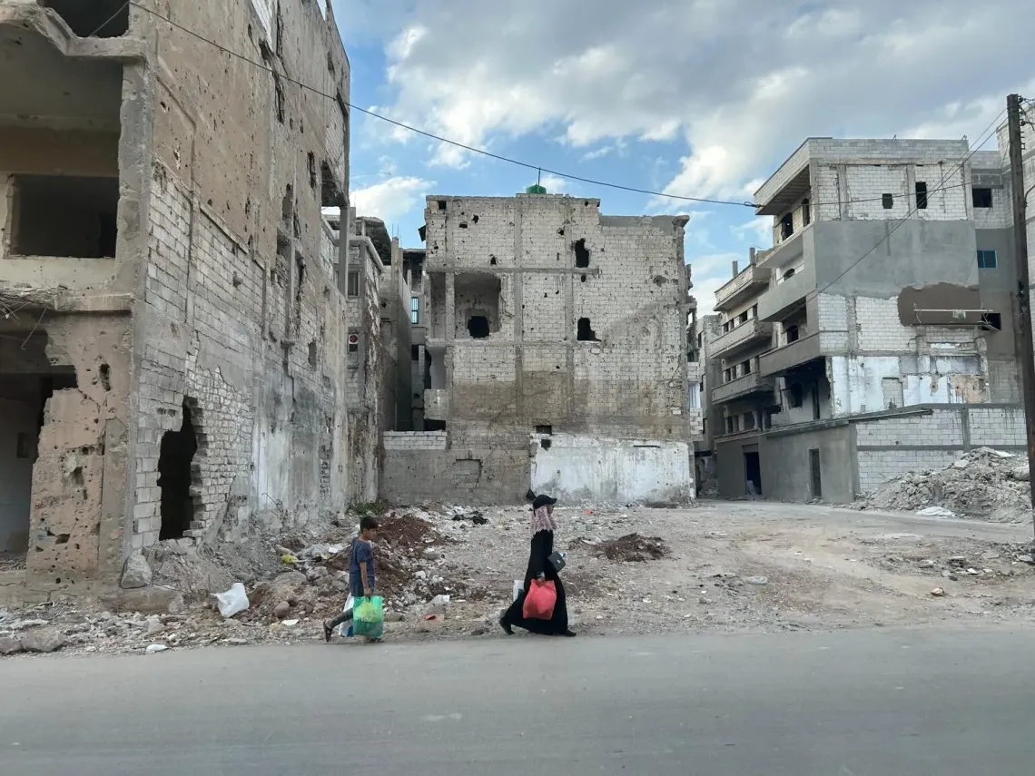Syria. A woman and a child walk past destroyed buildings.