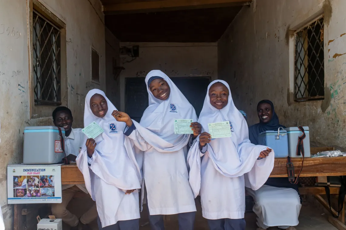 Three school girls in white head scarfs smile while posing for a picture in the hallway of their school.