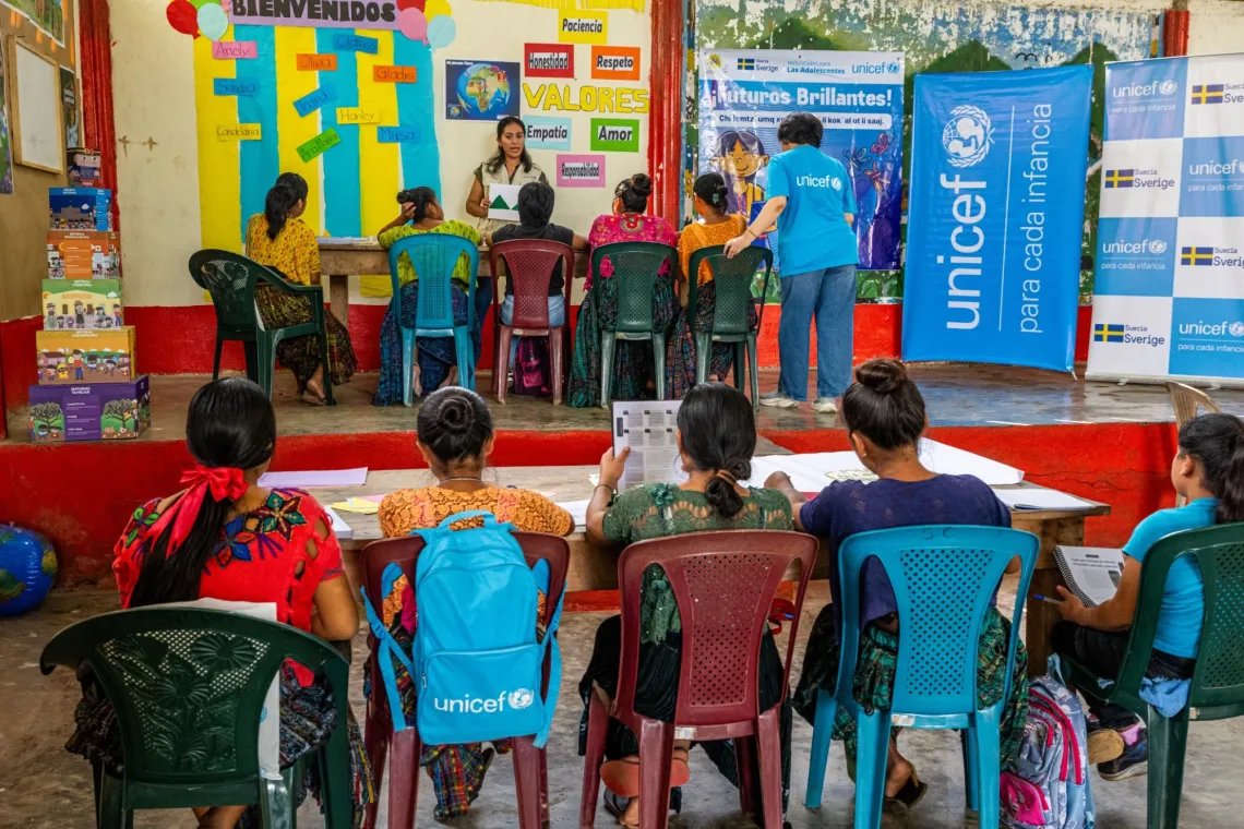 A group of children sit in a row on a stage with a UNICEF banner to the side, with a row of children looking on.