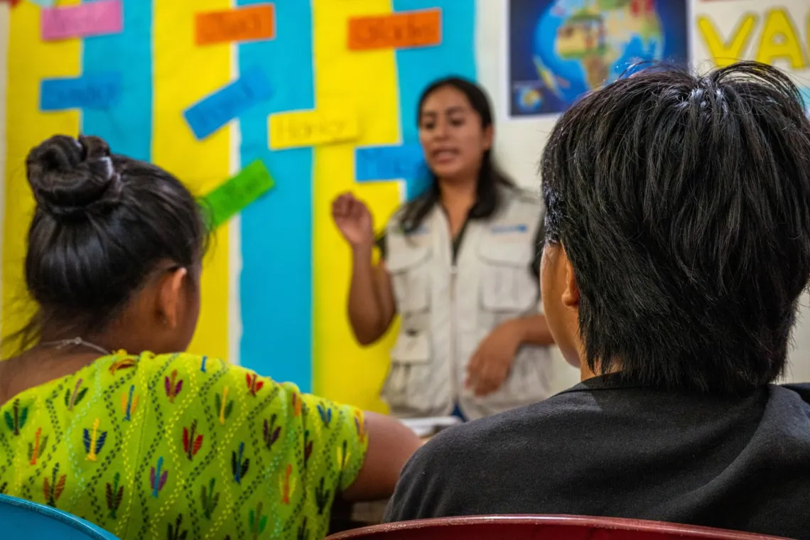A woman wearing a UNICEF waistcoat speaks to a group of young girls sitting in front of her.