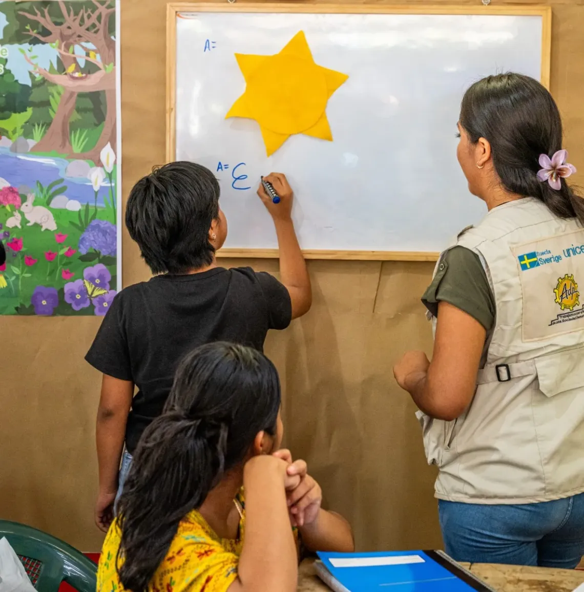 A young girl with short, cropped hair writes on a whiteboard, with a woman wearing a jacket with the UNICEF logo on the back stands next to her.