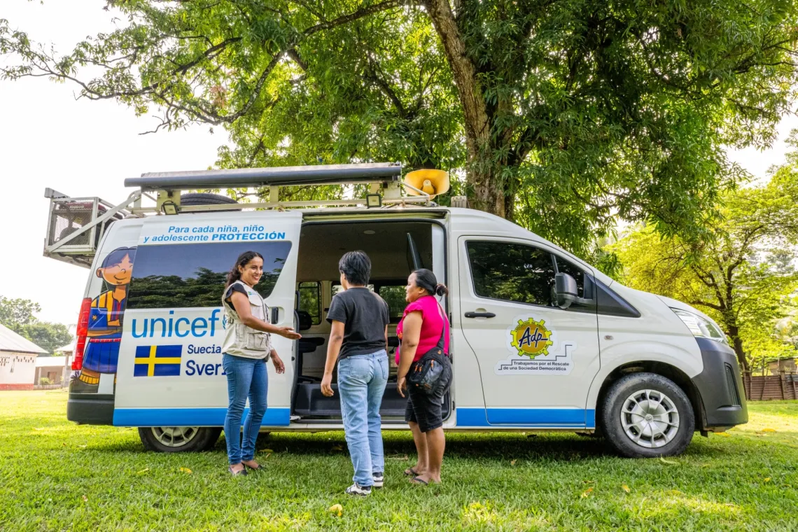 A mother and daughter are met by a woman wearing a UNICEF jacket in front of a van converted into a mobile unit.