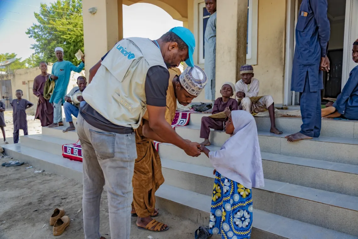 An immunization officer in a UNICEF vest holds the hand of a young girl in a white head scarf.
