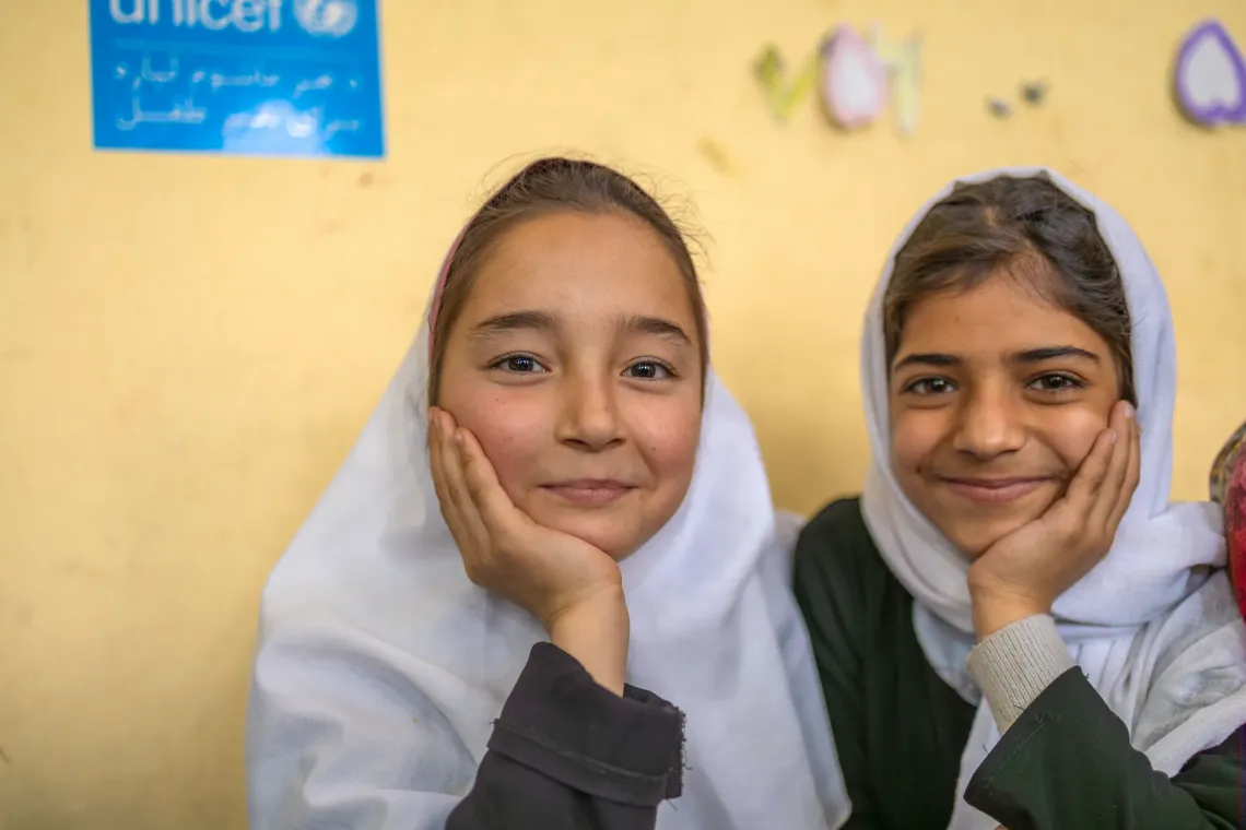 Two girls in Afghanistan rest their chins on their palms while smiling. 