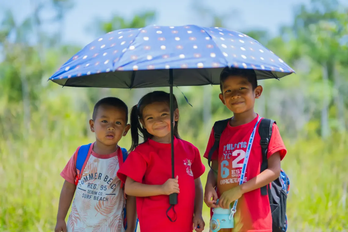 Three children stand smiling under a blue umbrella with polka dots. 