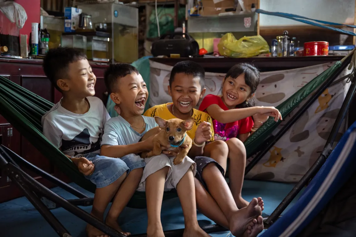 Four children, one holding a small dog, laugh while seated together in a hammock. 