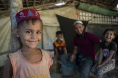 A young smiling boy looks into camera in a Rohingya refugee camp, as his family looks on in the background
