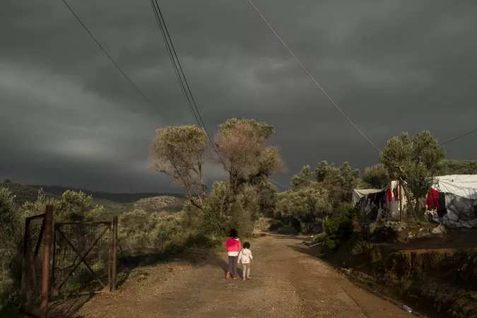 Two children walk on a dirt road surrounded by trees.