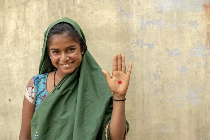 A girl smiles and holds her hand up, which has a red dot on it.