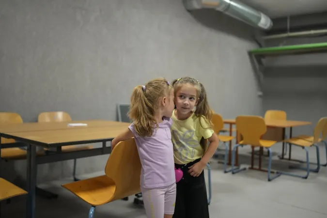 Two young girls talk to each other, standing in a classroom in front of school desks. 