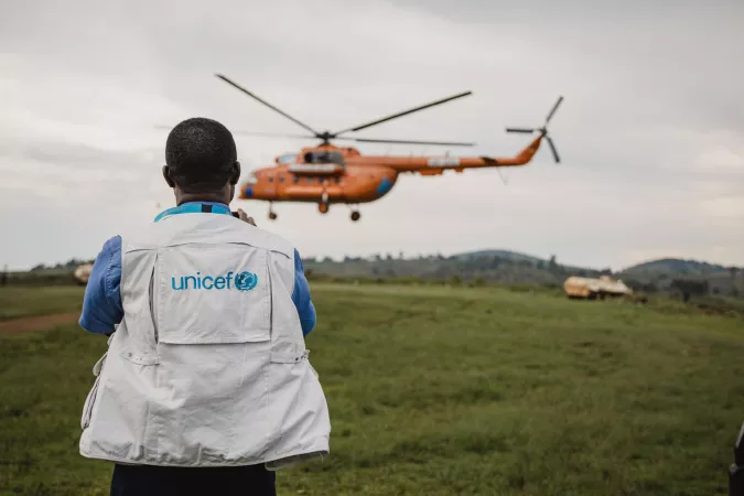 A UNICEF staff member stands in a green field as a helicopter lands.