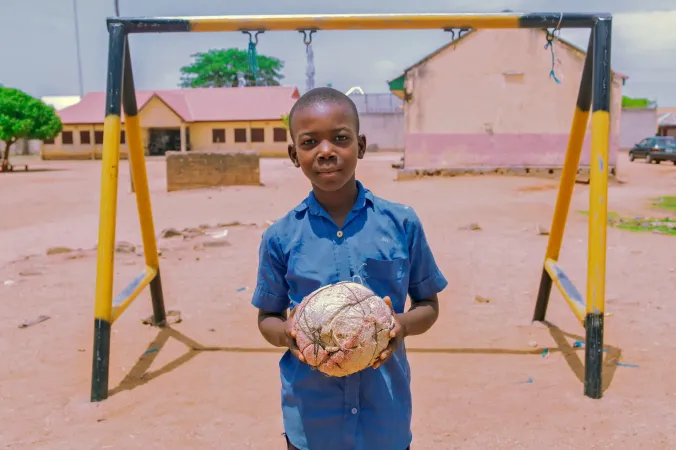 A child holds a football in front of a ne in a sandy field.