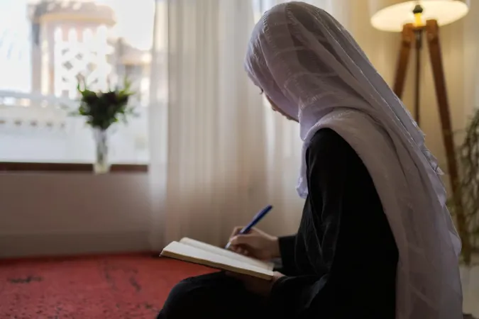 Girl wearing headscarf writes in book.