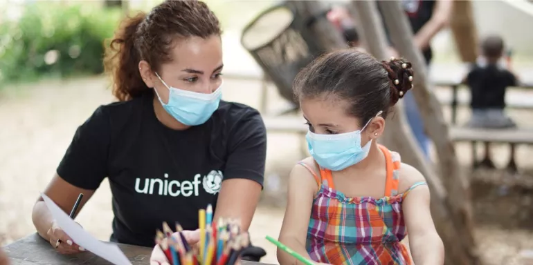 Woman in UNICEF-branded black t-shirt sits with a girl, both wearing face masks