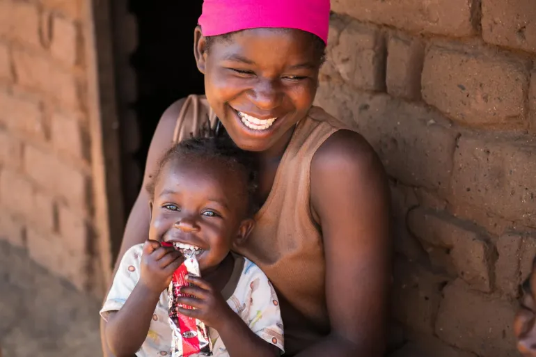 Child eats RUTF while sitting on mom's lap. Both are smiling. 
