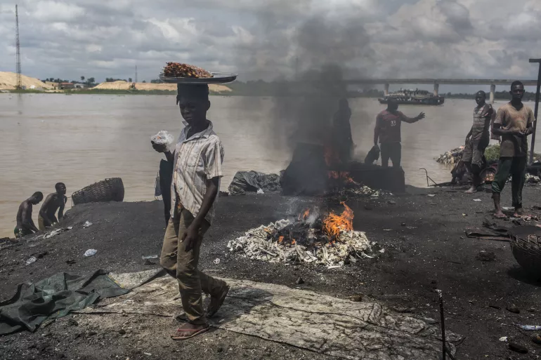 On 24 October 2016 in Yenagoa, Bayelsa State, Nigeria, children pass in front of a flame fed by waste and rubber materials in order to make Kanda, a type of smoked meat, at an abattoir. The workers at the slaughterhouse use cow bones, rubber tires, electric wires, aluminum cans and other waste to sustain the flames, making the fumes very dangerous to inhale.