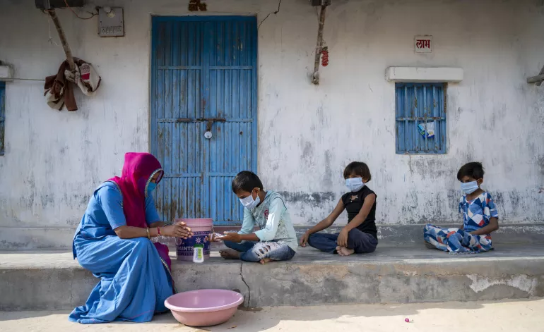 A mother and her children wash their hands outside their home using a bucket, water, and soap.