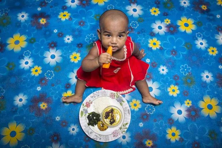 Child sits and eats nutritious plate of food, looking at camera.