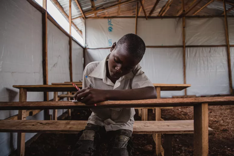 A child sits at a desk and writes in tented classroom.